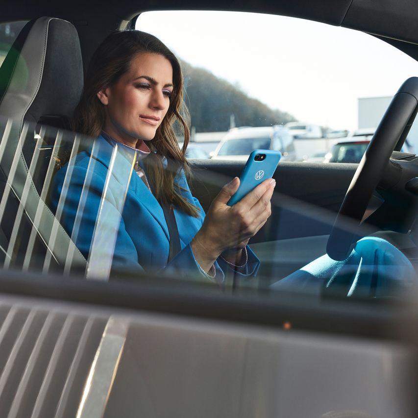 Eine in blau gekleidete Frau sitzt in einem geparkten Volkswagen mit einem Smartphone in ihrer Hand.