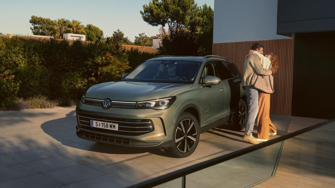 Grüner VW Tiguan parkt in Frontansicht auf der Einfahrt eines modernen Wohnhauses. Ein junges Paar steht daneben und umarmt sich.