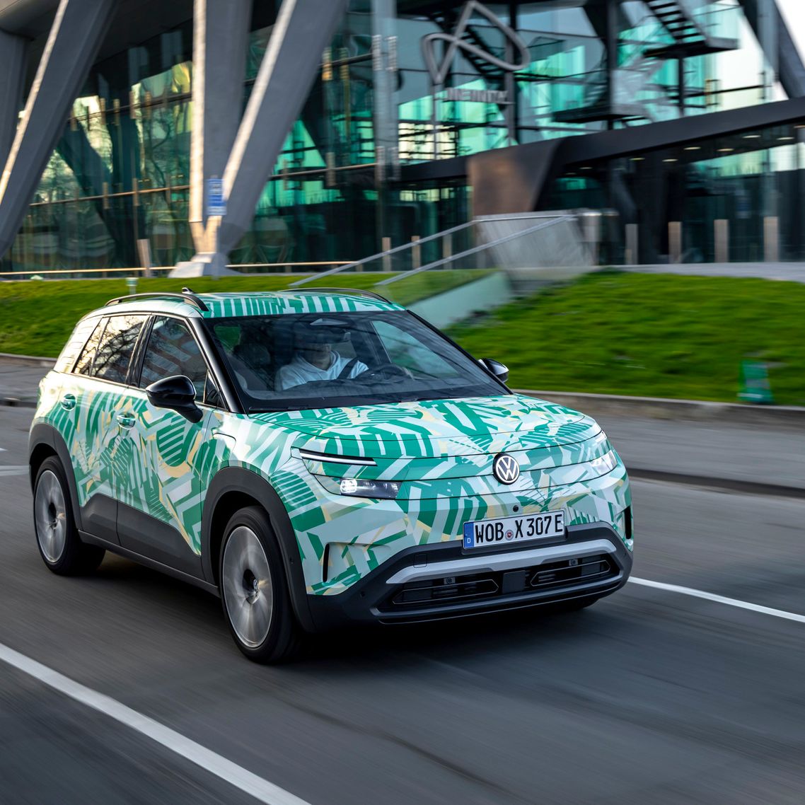 Ein Auto, das mit grünen und weißen Mustern bedeckt ist, fährt auf einer Straße vor einem modernen Gebäude mit großen Glasfenstern und einer auffälligen Architektur. Im Hintergrund sind grüne Grasflächen und Treppen zu sehen. Das Fahrzeug scheint in Bewegung zu sein.