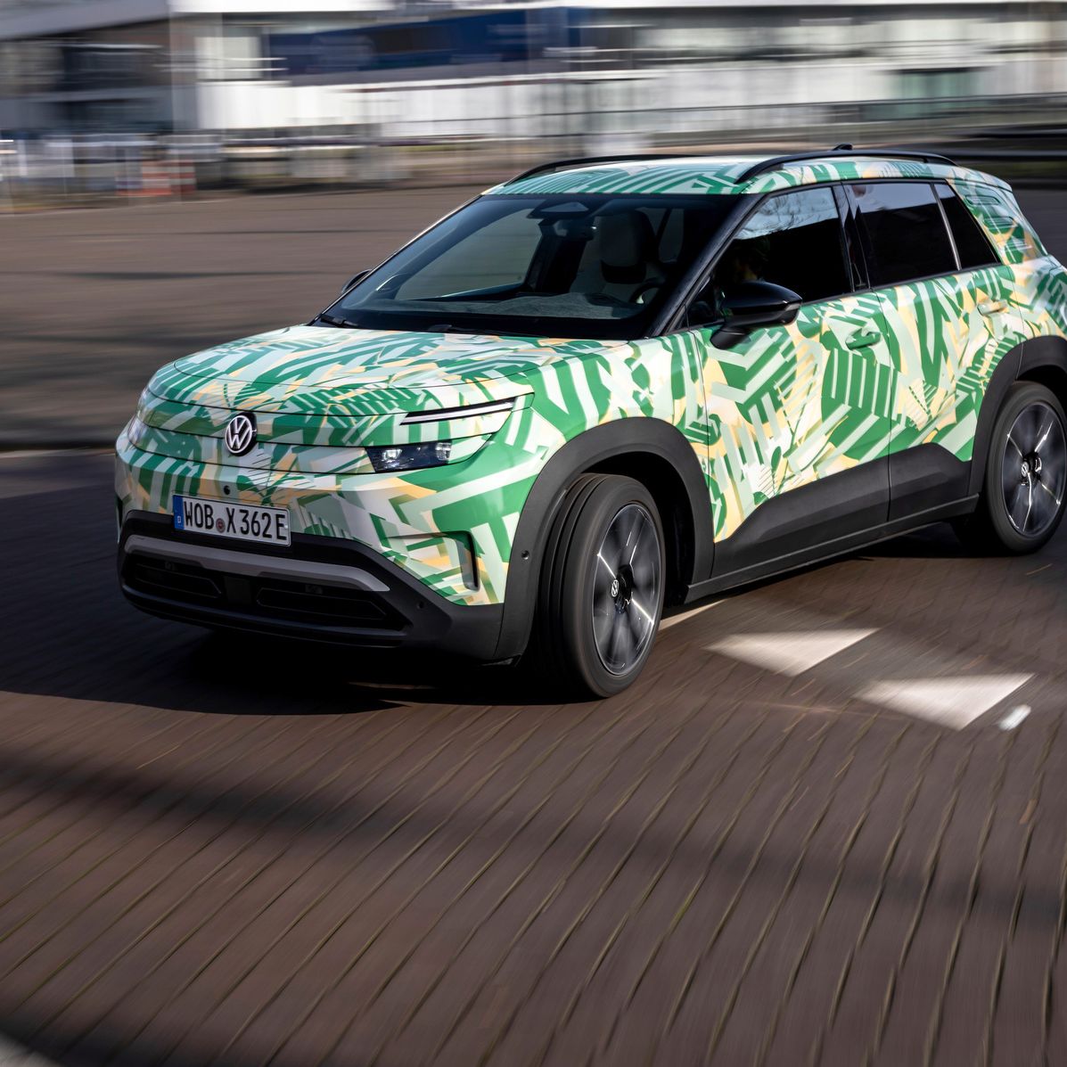 Ein Auto mit grüner und gelber Tarnlackierung fährt eine Straße entlang. Es handelt sich um einen SUV mit einem Kennzeichen, das "WOB PA 342E" zeigt. Im Hintergrund sind unscharfe Gebäude zu sehen.