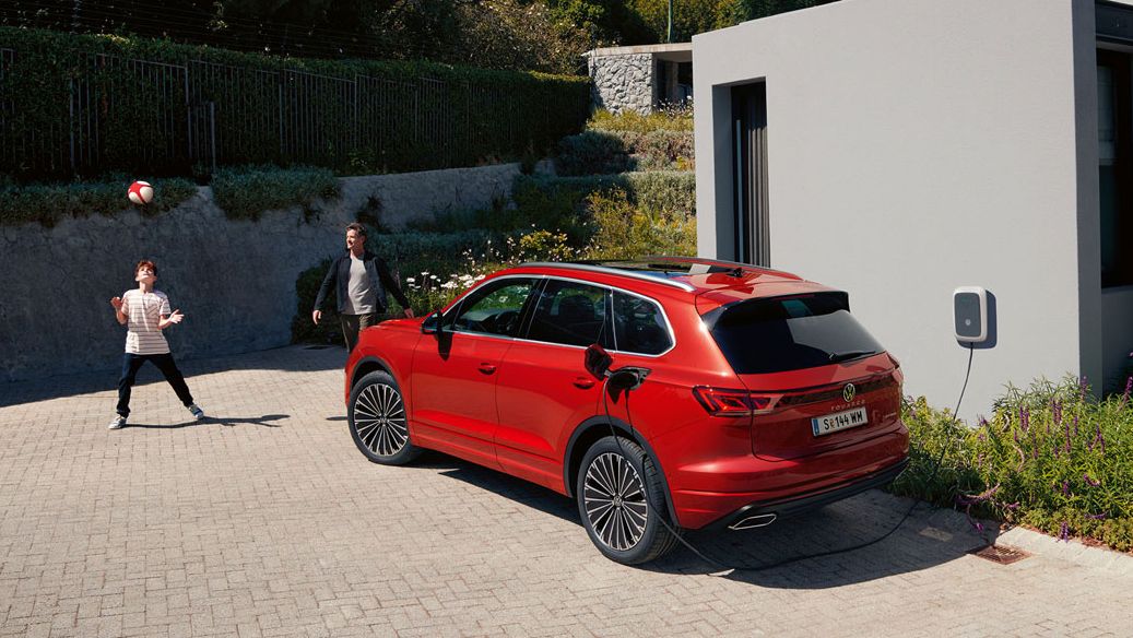 Ein VW Touareg Elegance eHybrid, Ansicht von schräg hinten, an einer Ladesäule vor einem modernen Haus.