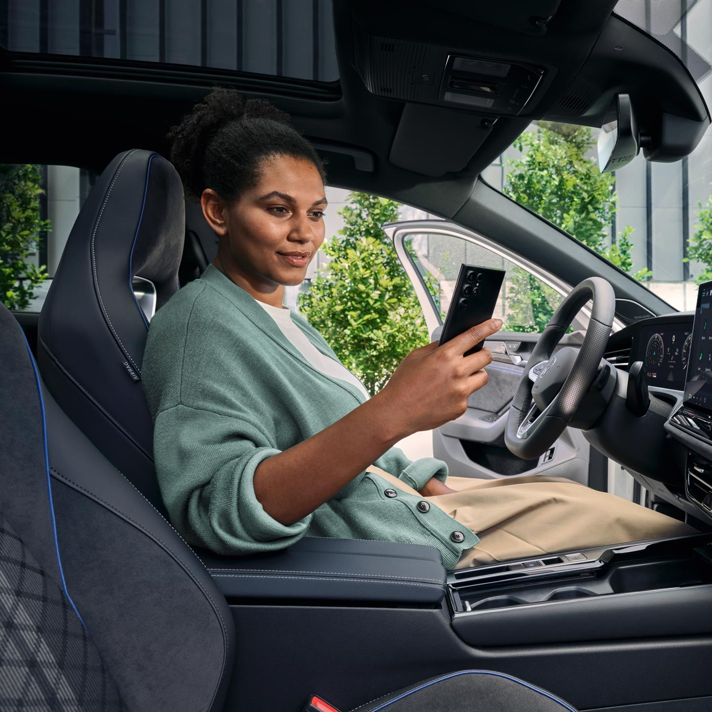 Eine Frau sitzt bei geöffneter Tür auf dem Fahrersitz des geparkten VW Passat und blickt auf ihr Smartphone.