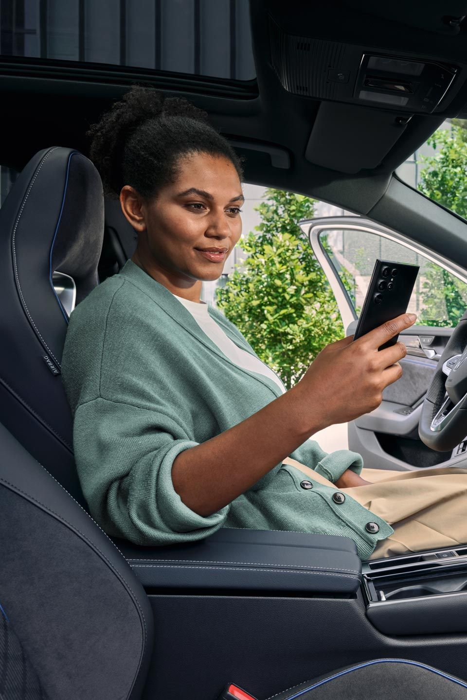 Eine Frau sitzt bei geöffneter Tür auf dem Fahrersitz des geparkten VW Passat und blickt auf ihr Smartphone.
