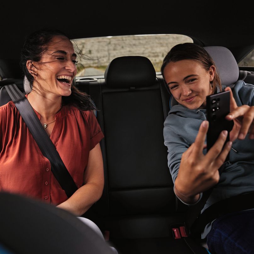 2 Peronen sitzen auf der Rückbank eines Volkswagen und schauen lächelnd auf ein Mobiltelefon