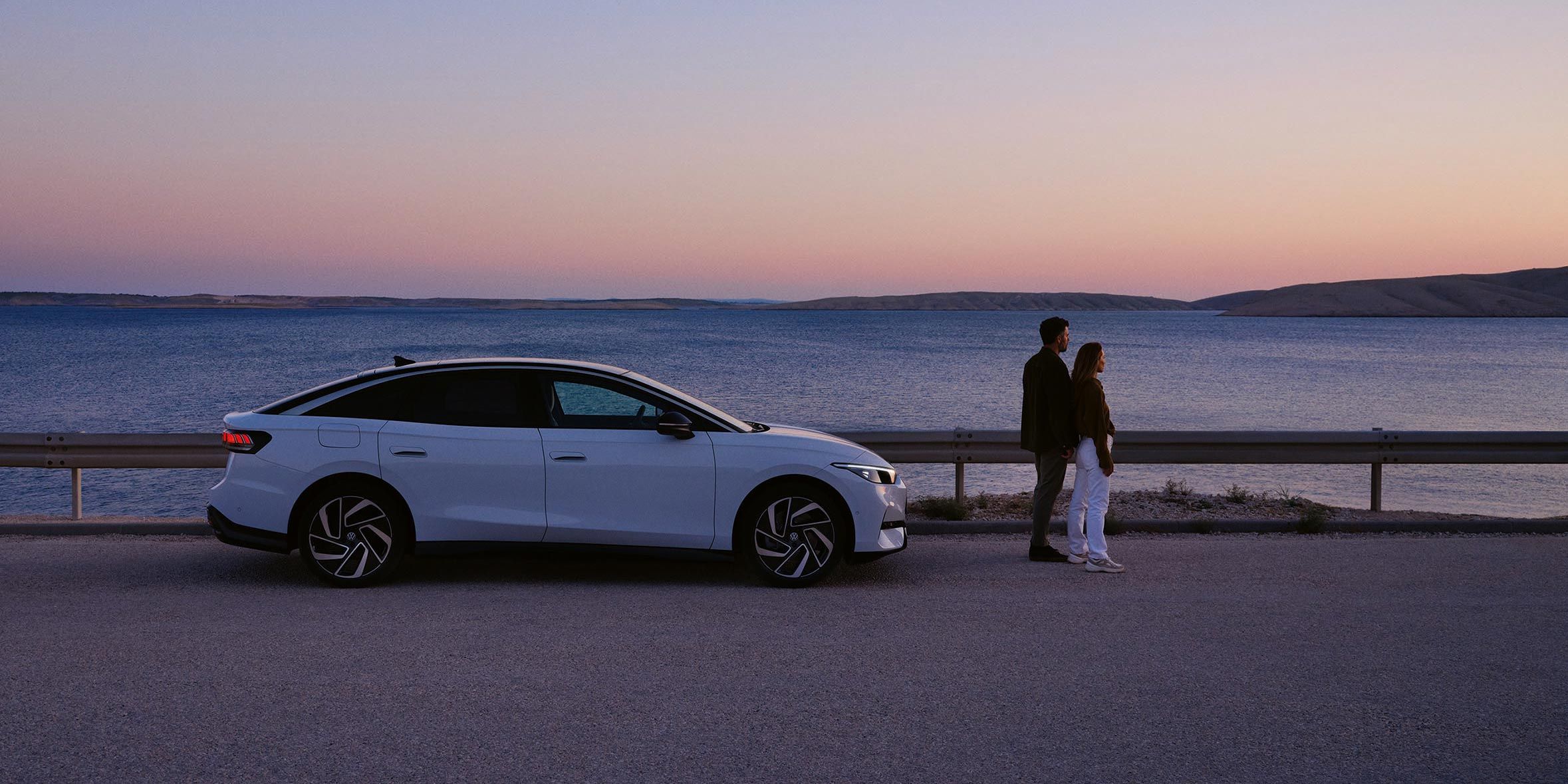 Ein VW ID.7 steht geparkt am Meer. Vor dem Auto steht ein Pärchen, das in den Sonnenuntergang blickt.