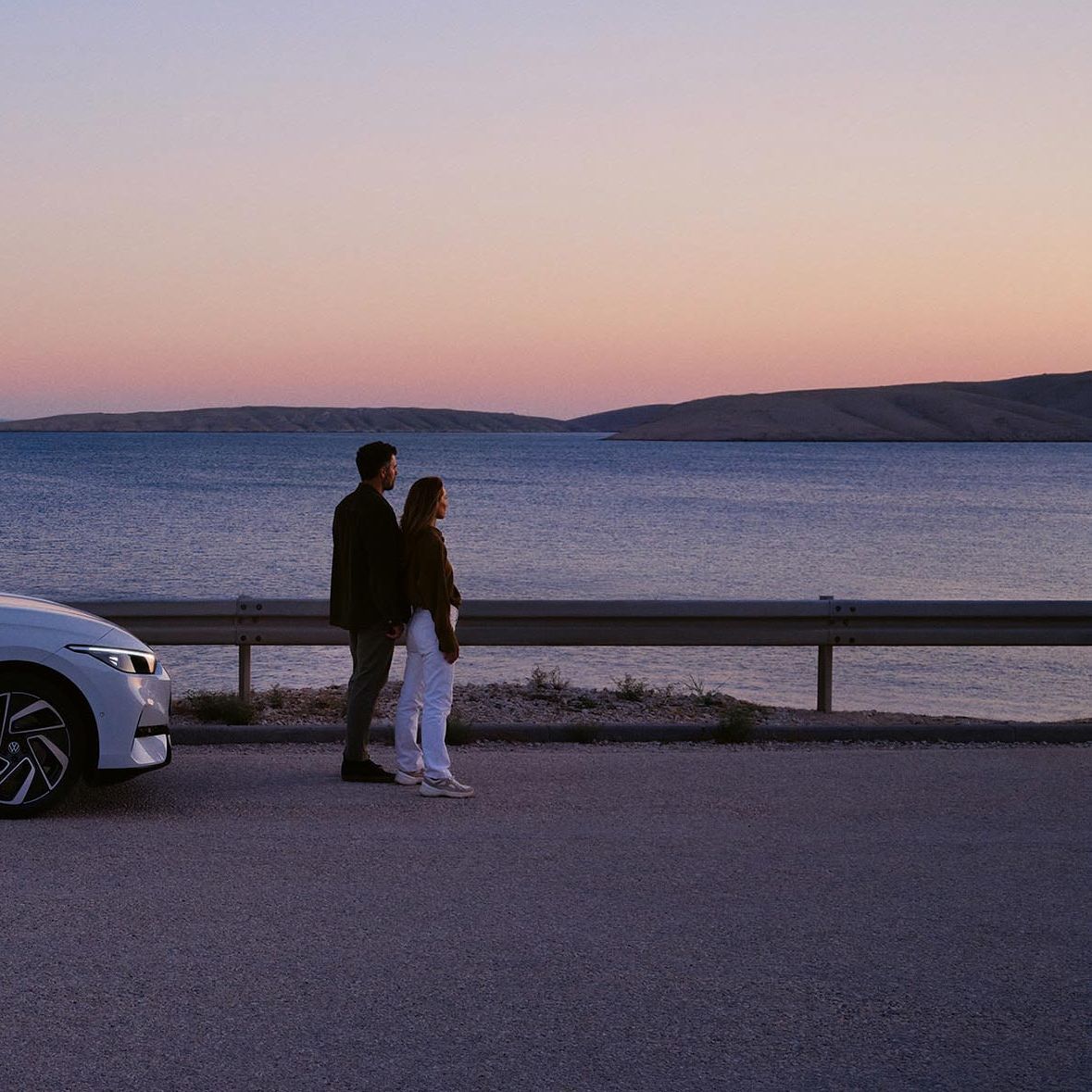 Ein VW ID.7 steht geparkt am Meer. Vor dem Auto steht ein Pärchen, das in den Sonnenuntergang blickt.