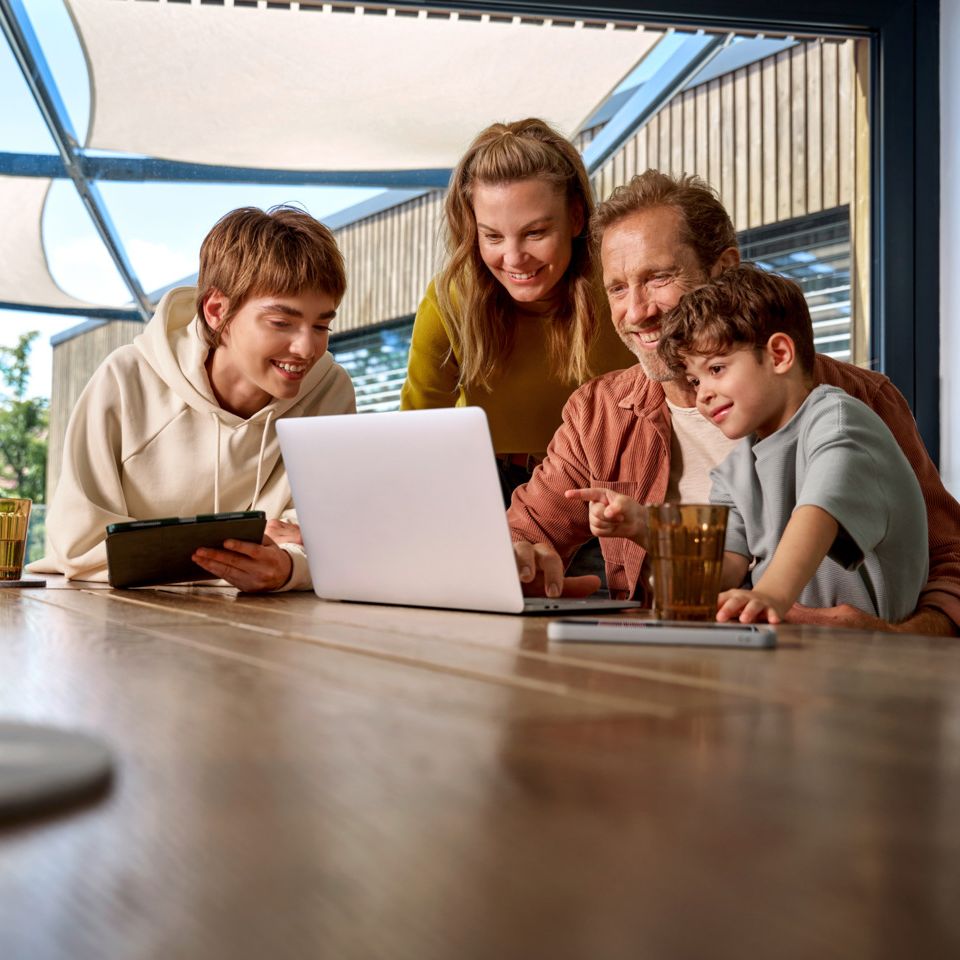 Eine Familie ist um einen Tisch versammelt und schaut fröhlich auf einen Laptop-Bildschirm
