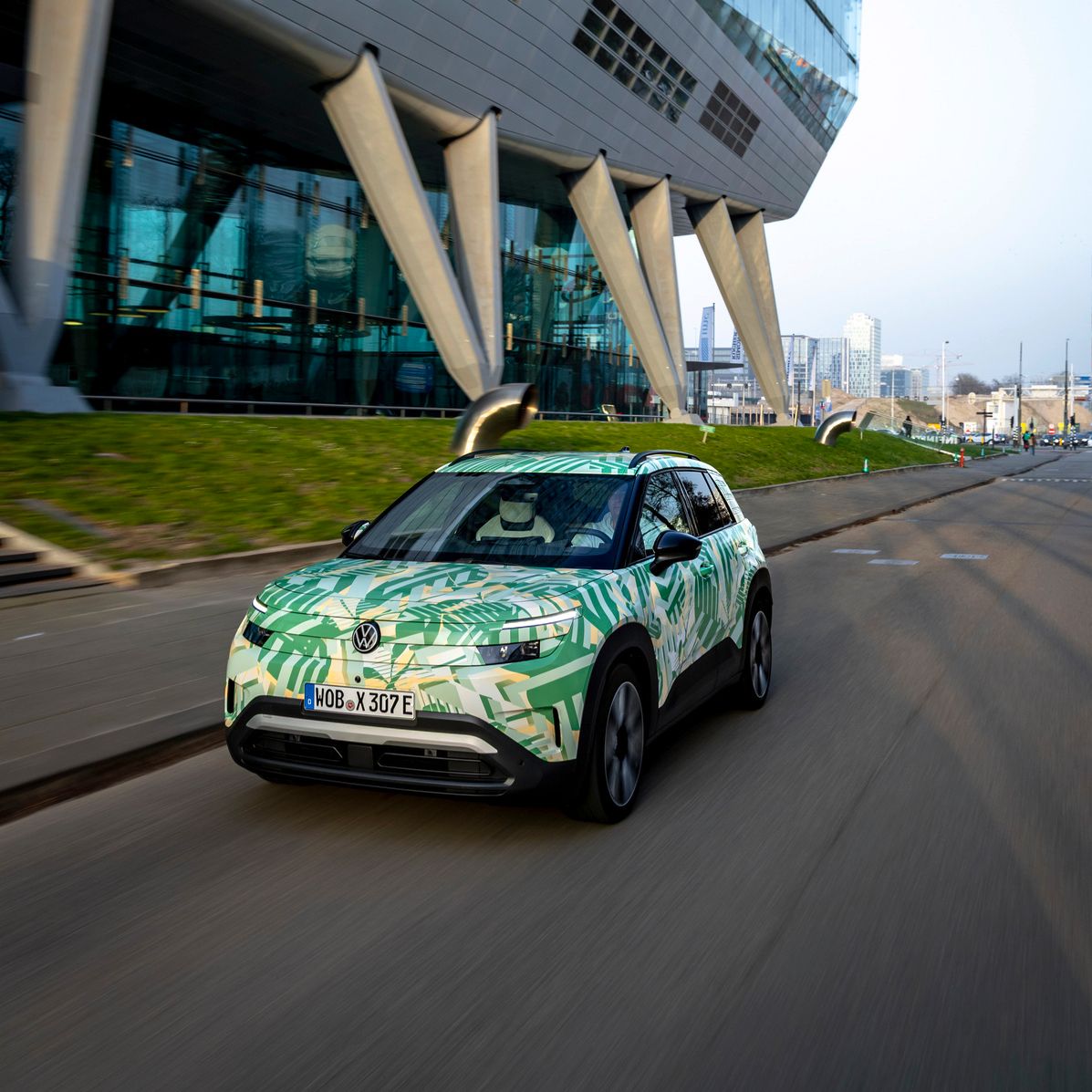 Ein Elektroauto fährt auf einer Straße neben einem modernen Gebäude mit schrägen Stützen und großer Glasfassade. Das Auto hat eine grüne und weiße Tarnlackierung. Im Hintergrund sind weitere Gebäude und eine graue, bewölkte Skyline zu sehen.