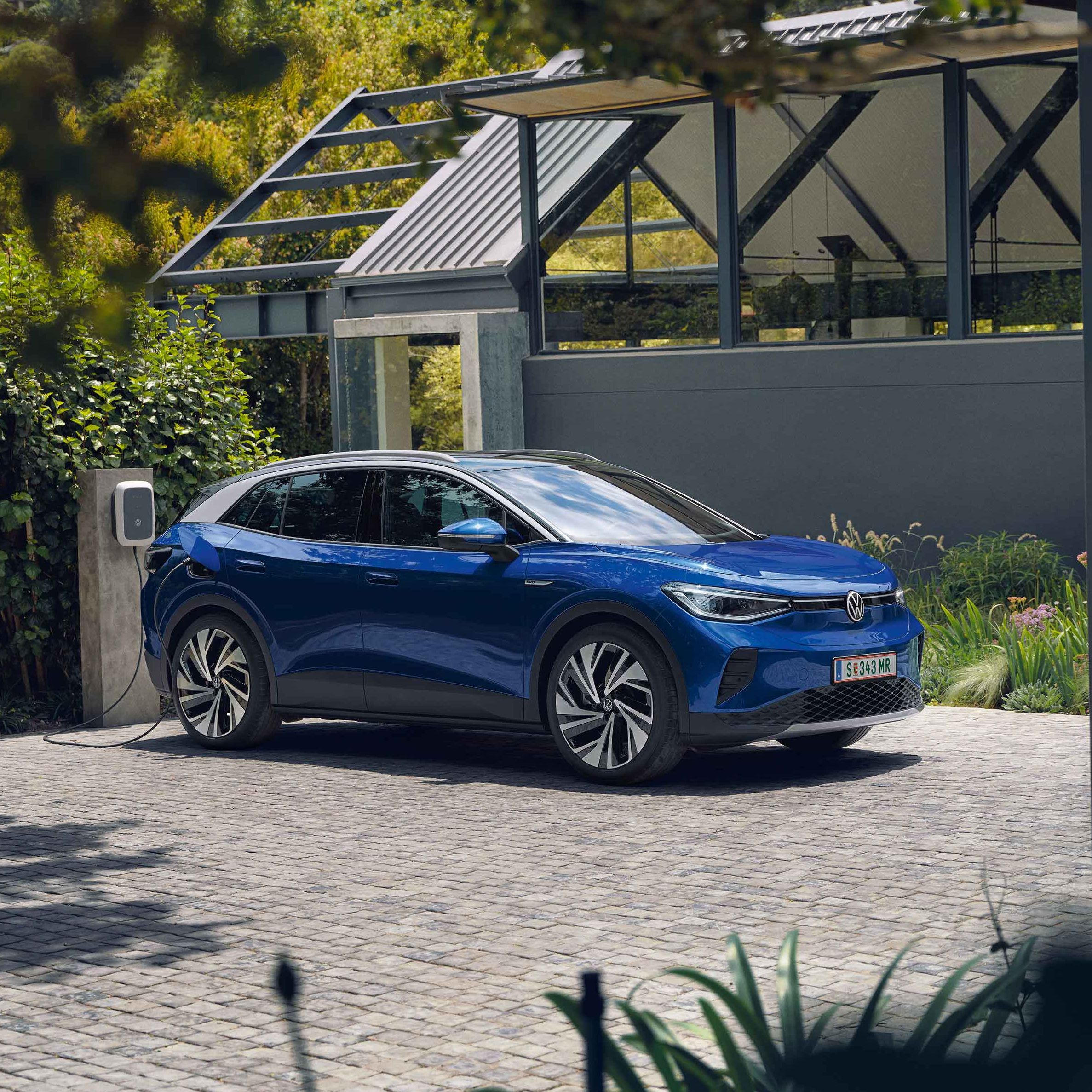 Ein blauer VW ID.4 parkt seitlich vor einem grauen, modernen Haus und lädt an der ID. Charger Wallbox. Im Hintergrund viel grün.