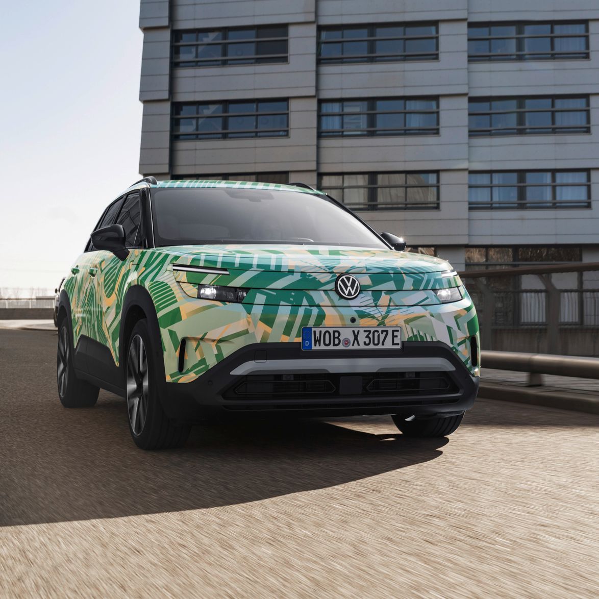 Ein ID. Cross von Volkswagen mit einem auffälligen grünen und gelben Camouflage-Design fährt auf einer städtischen Straße. Im Hintergrund sind moderne Gebäude zu sehen. Das Nummernschild des Autos beginnt mit "WOB".