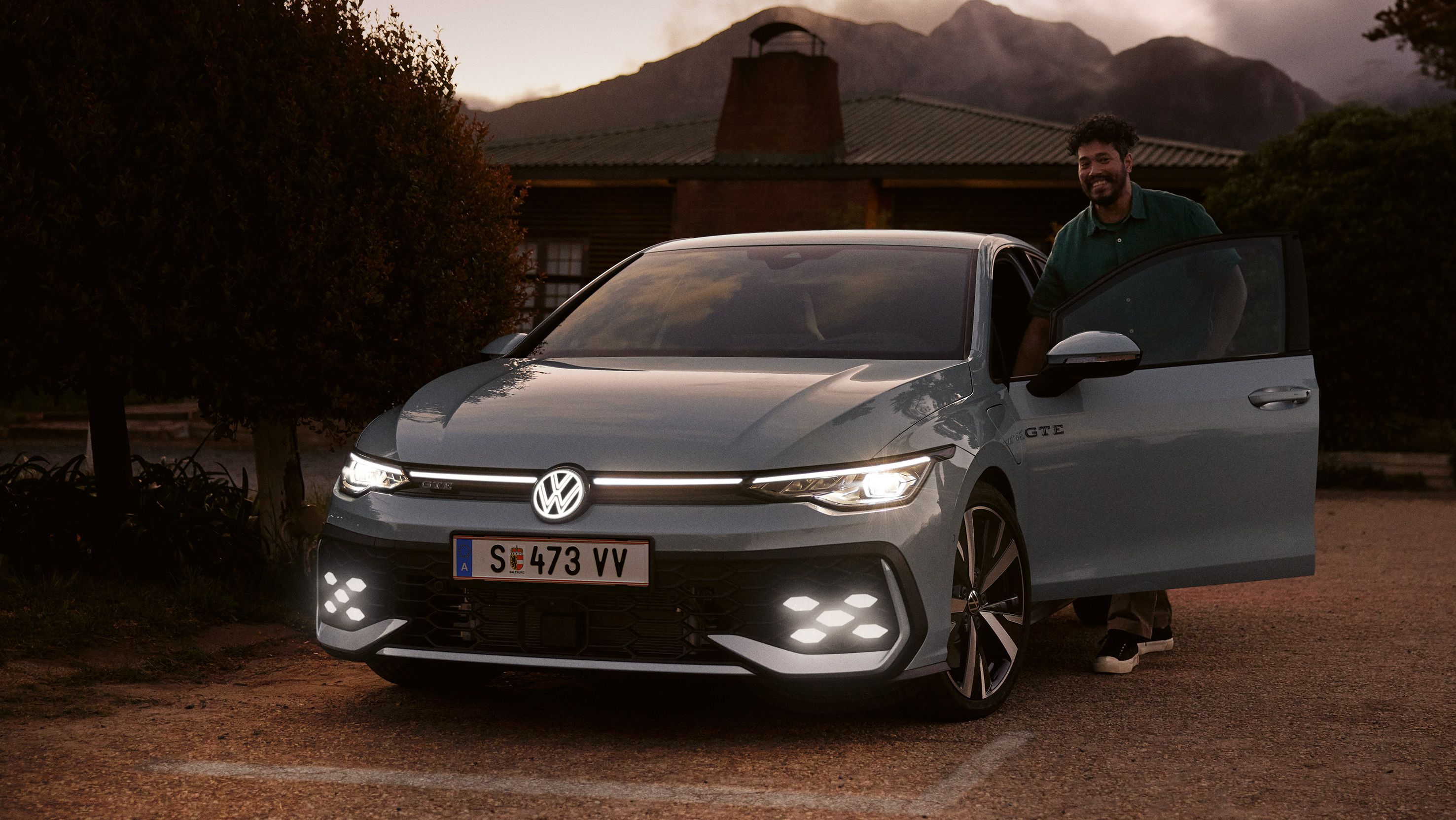 Frontansicht eines grauen VW Golf GTE mit IQ. Light LED Matrix Scheinwerfern in abendlicher Stimmung, Mann steigt in Golf GTE