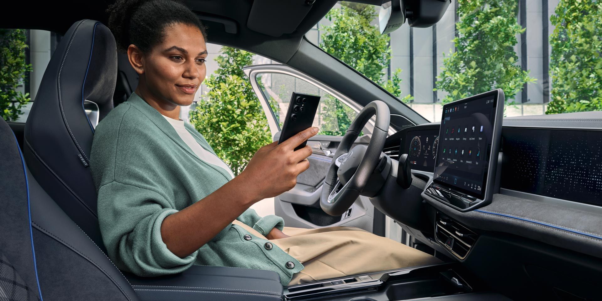 Eine Frau sitzt bei geöffneter Tür auf dem Fahrersitz des geparkten VW Passat und blickt auf ihr Smartphone.