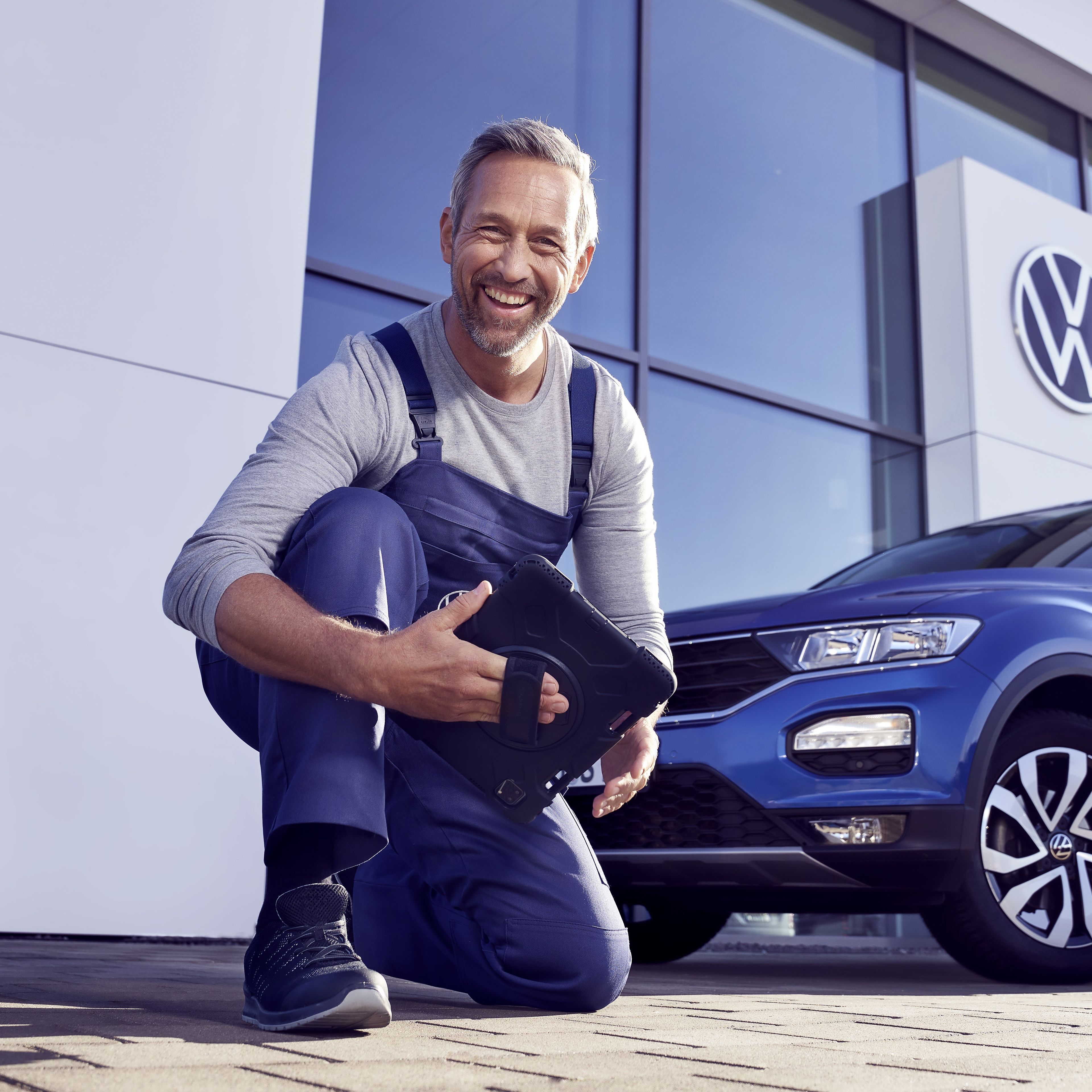 Ein Volkswagen Service Techniker mit Tablet, kniet vor einem blauen Volkswagen T-Roc