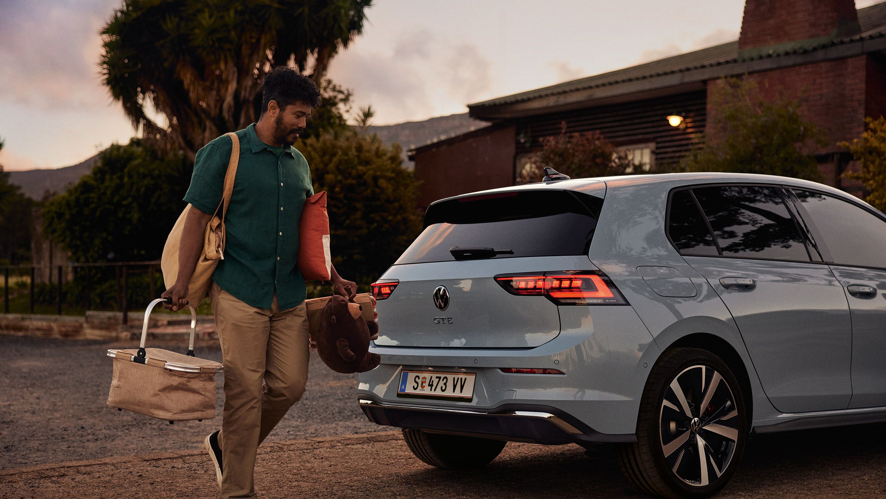 Heckansicht eines grau-blauen VW Golf GTE in abendlicher Stimmung, Mann trägt einen Korb und Spielsachen