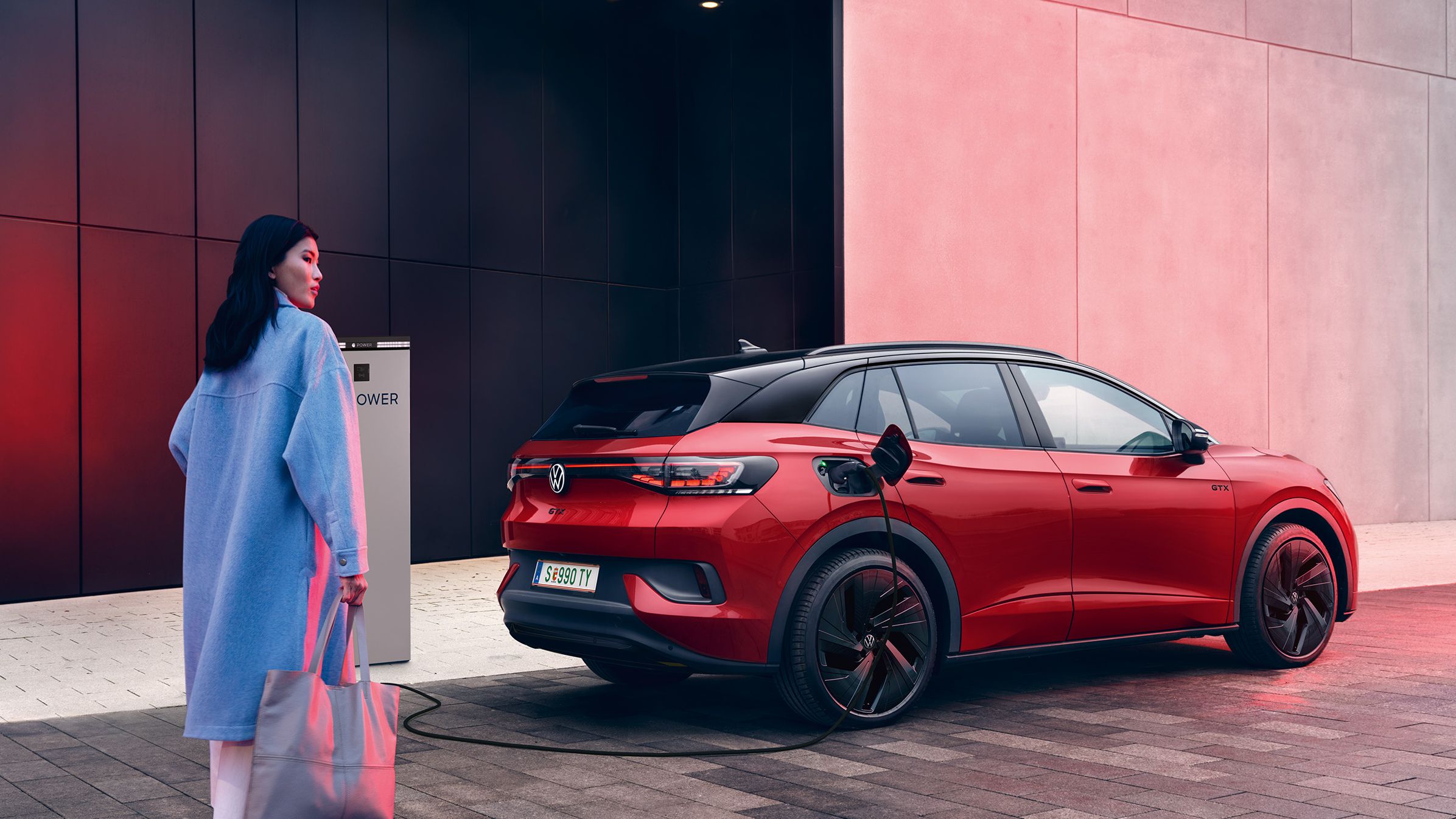 Heck- und Seitenansicht eines roten VW ID.4 GTX der an Ladesäule geladen wird, Frau geht auf Fahrzeug zu