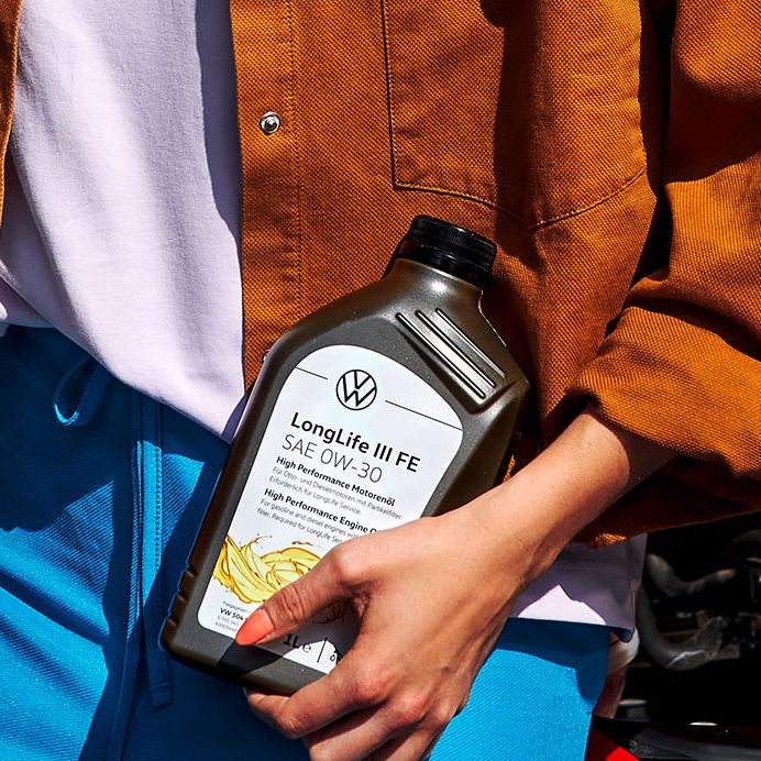 Eine Frau mit einer Flasche Motoröl in der Hand lehnt an einem roten VW mit geöffneter Motorhaube