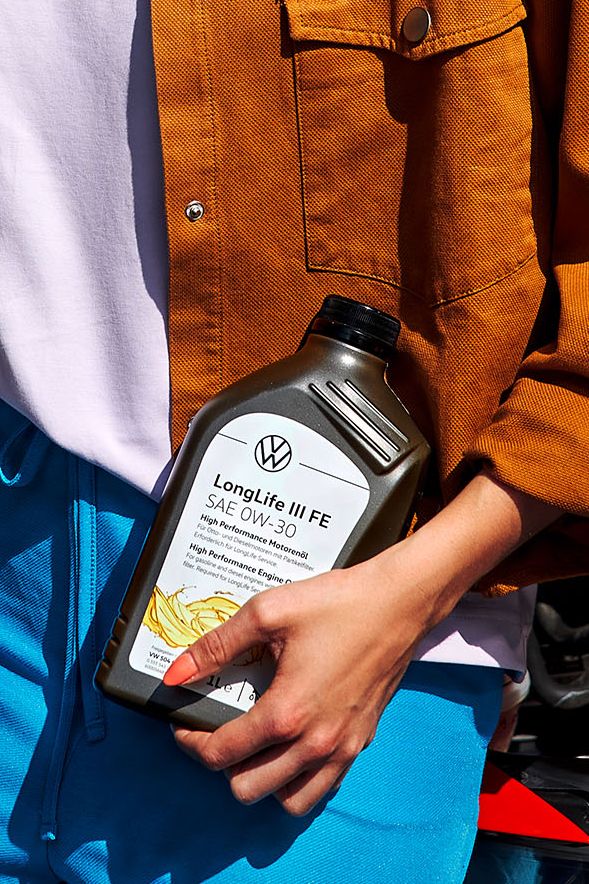 Eine Frau mit einer Flasche Motoröl in der Hand lehnt an einem roten VW mit geöffneter Motorhaube
