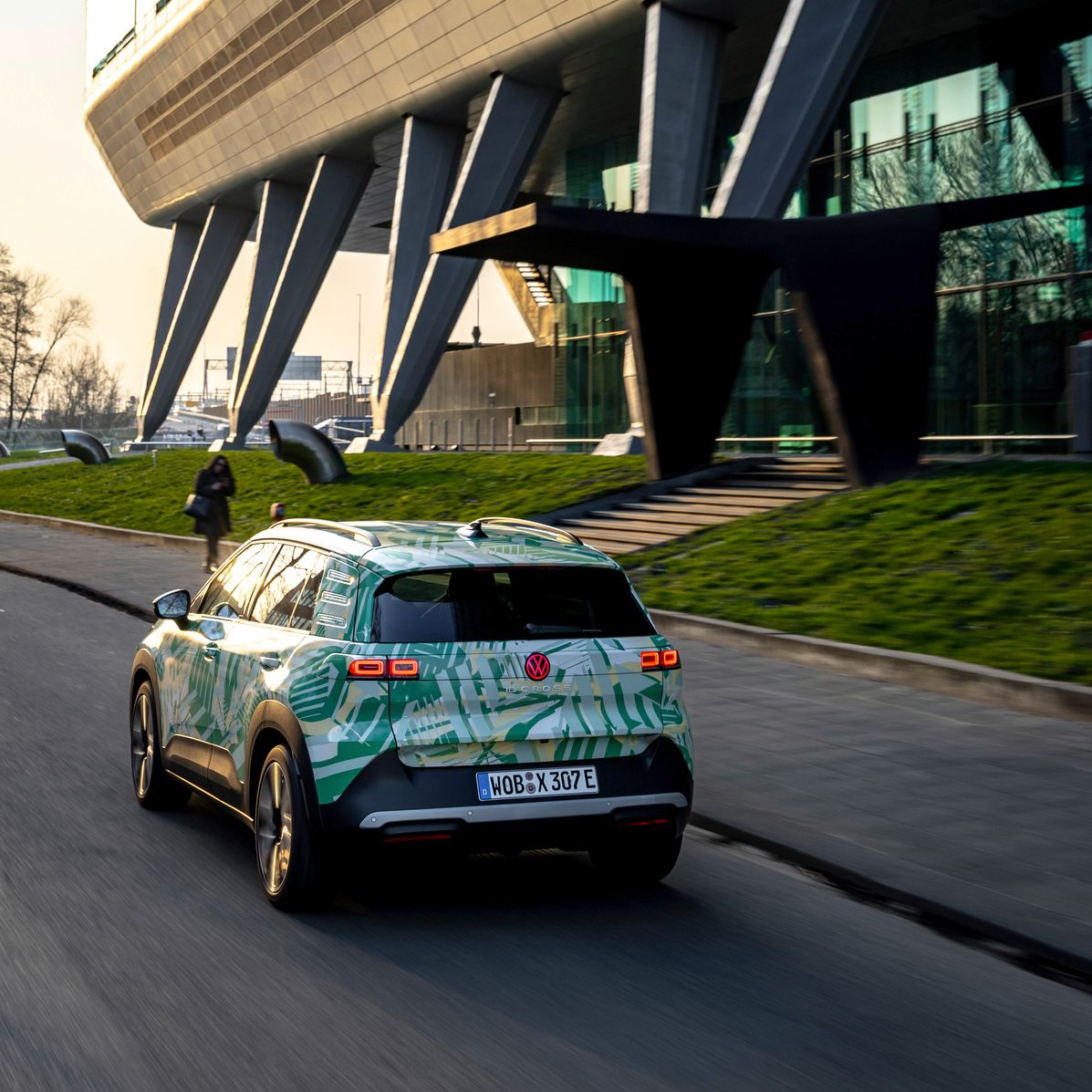 Ein Auto fährt auf einer Straße an einem modernen Gebäude mit Glasfassade und Stelzen vorbei. Das Auto hat eine auffällige, hellgrüne und gemusterte Lackierung. Im Hintergrund sind einige Bäume und eine Fußgängerin zu sehen.