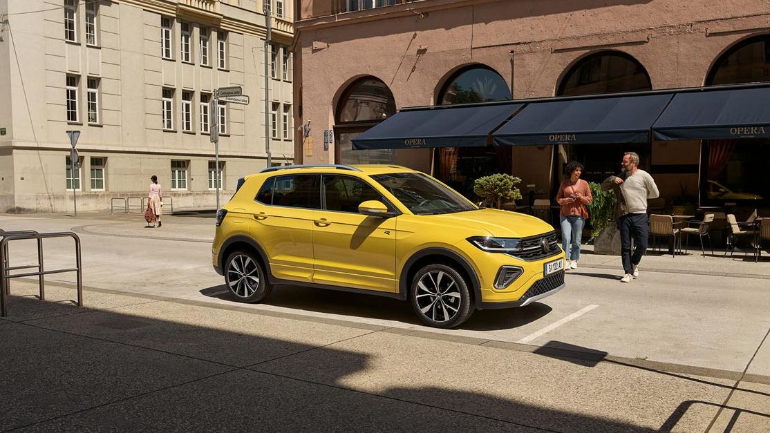 VW T-Cross in gelb parkt seitlich vor einem urbanen Straßencafé, ein Päarchen geht auf den Wagen zu.