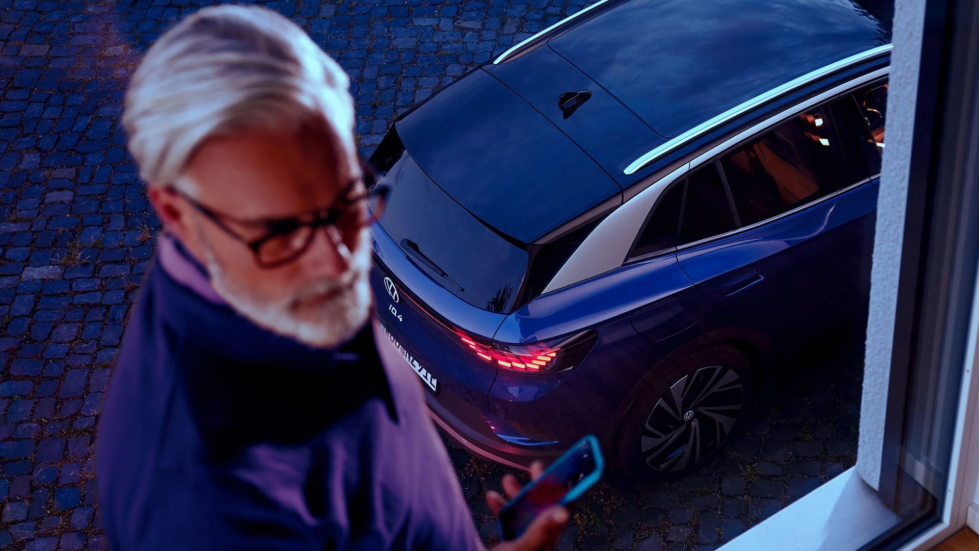 Mann mit Smartphone in der Hand steht am Fenster, im Hintergrund steht ein blauer VW ID.4