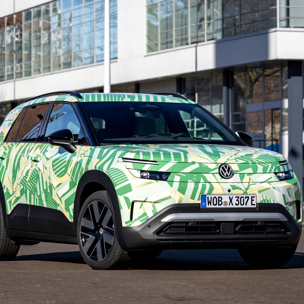 Ein Volkswagen Elektro-SUV steht vor modernen Gebäuden. Das farbenfrohe Fahrzeug hat ein grünes und cremefarbenes Camouflage-Muster mit Blattmotiven. Das Nummernschild zeigt eine Zulassung aus Wolfsburg.