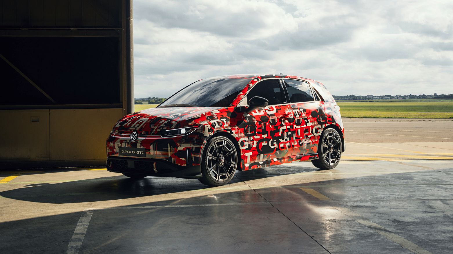 Ein ID. Polo GTI das mit Tarnmustern lackiert ist, steht in einer offenen Garage. Der Himmel im Hintergrund ist bewölkt. Die Lackierung könnte auf ein neues Modell oder eine Testversion hinweisen.