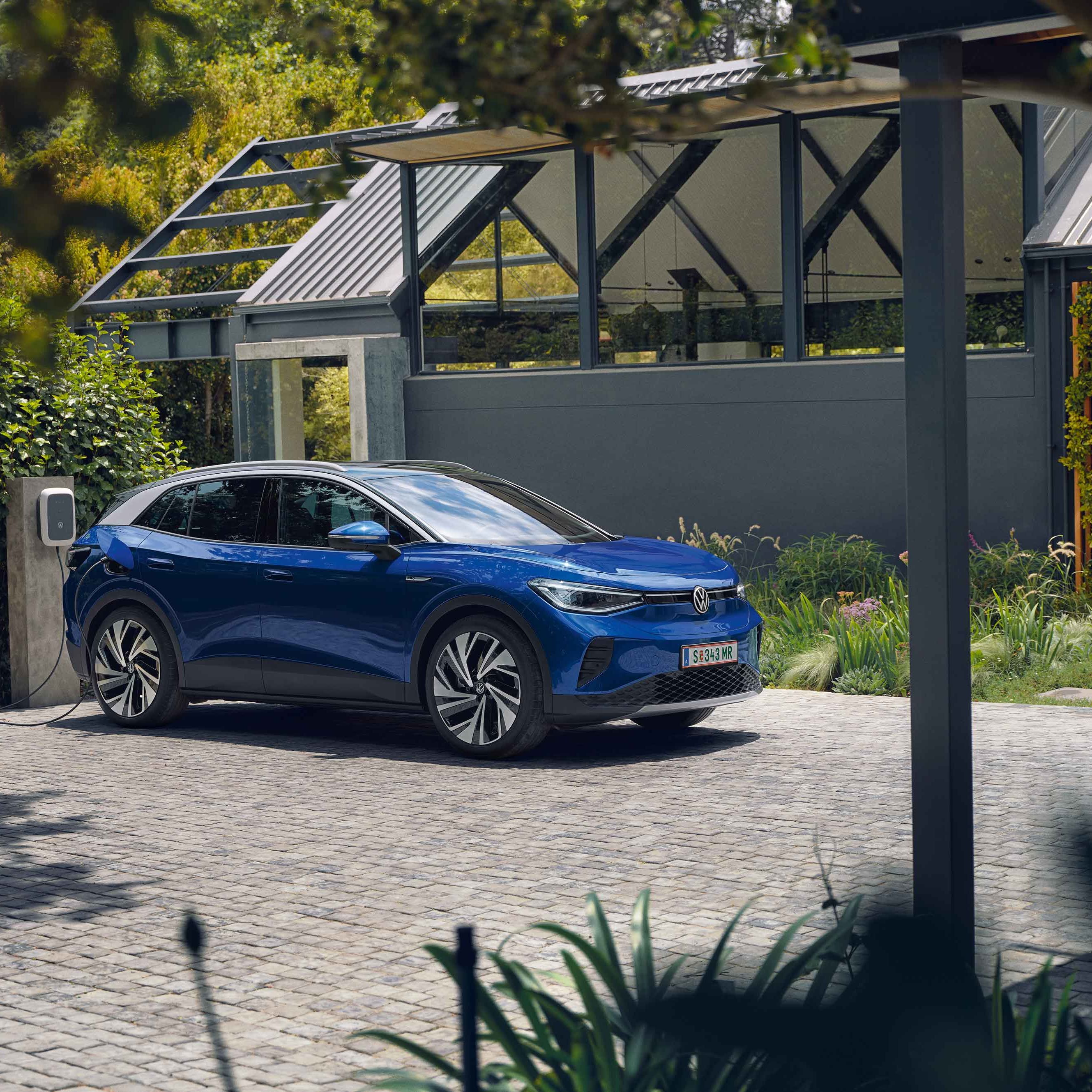 Ein blauer VW ID.4 parkt seitlich vor einem grauen, modernen Haus und lädt an der ID. Charger Wallbox. Im Hintergrund viel grün.
