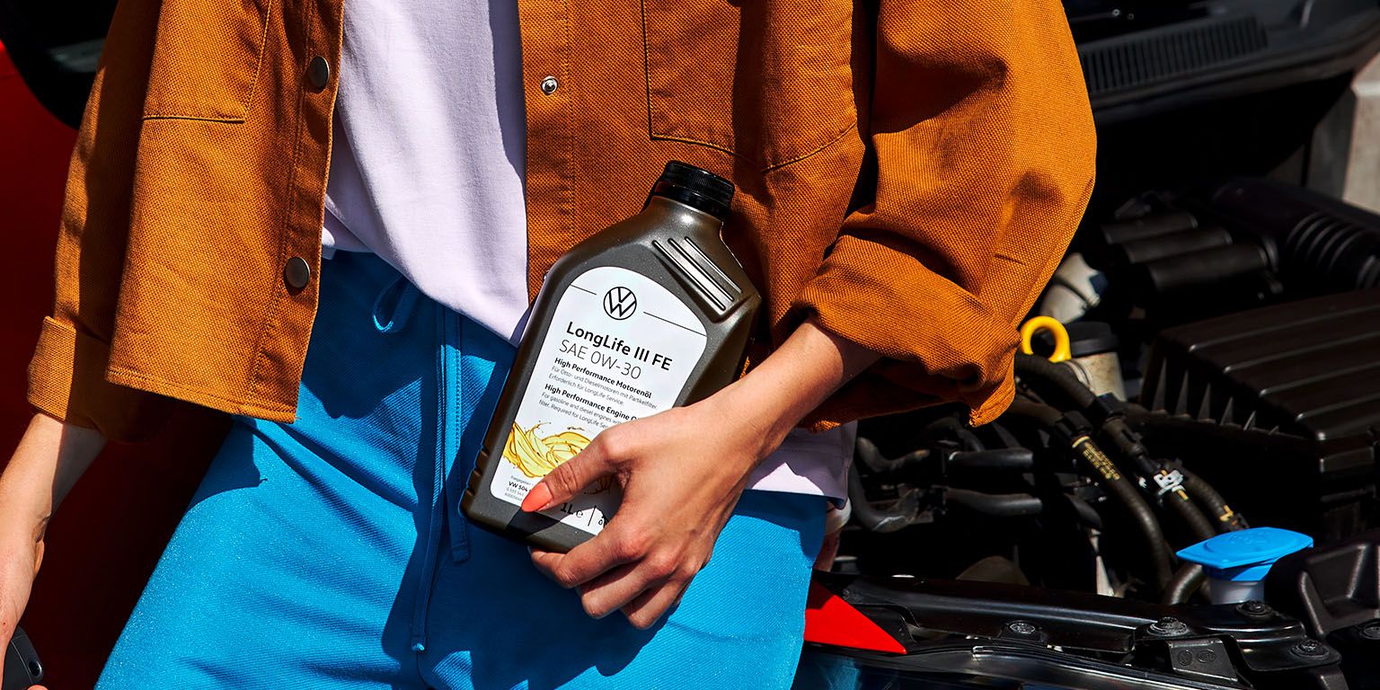 Eine Frau mit einer Flasche Motoröl in der Hand lehnt an einem roten VW mit geöffneter Motorhaube