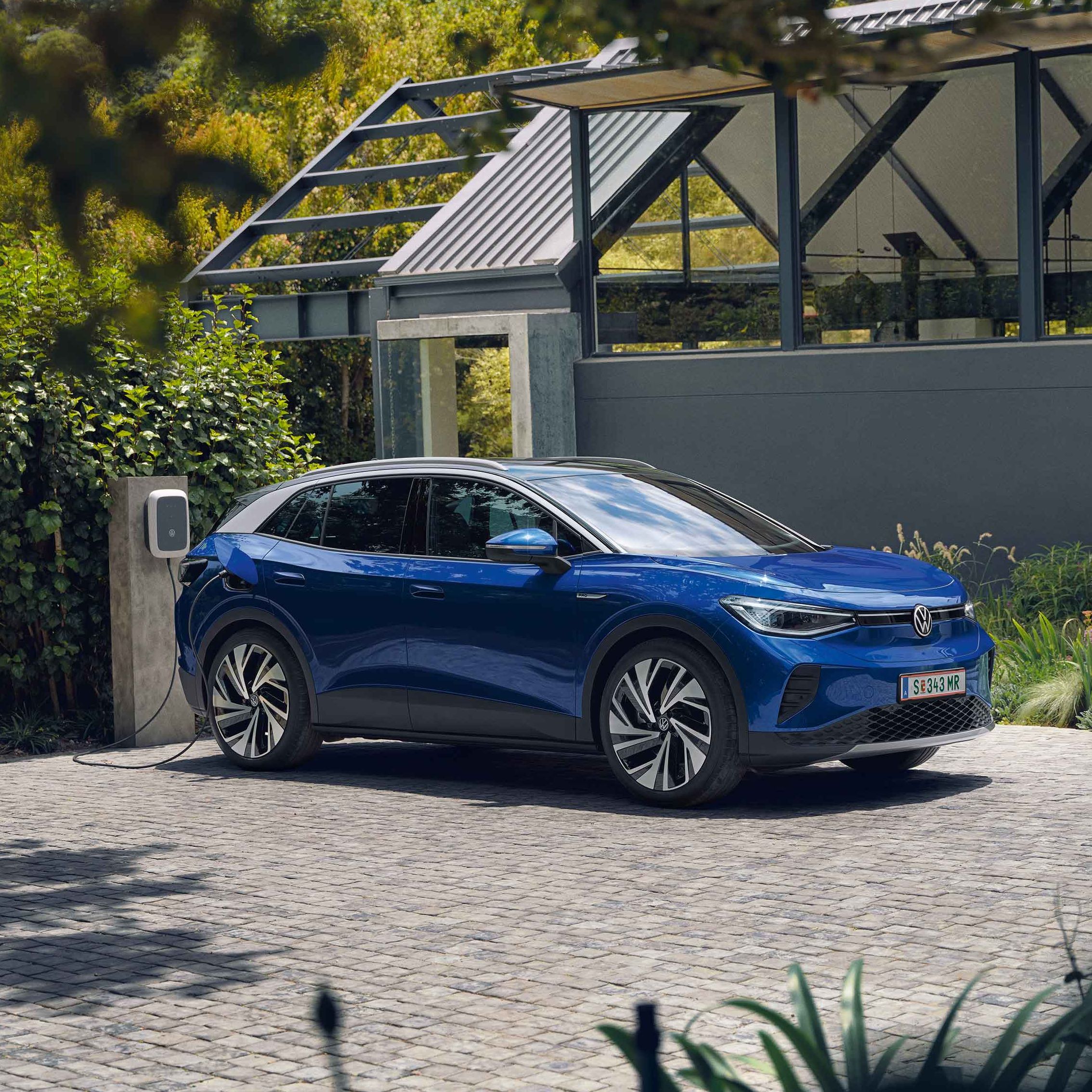 Ein blauer VW ID.4 parkt seitlich vor einem grauen, modernen Haus und lädt an der ID. Charger Wallbox. Im Hintergrund viel grün.