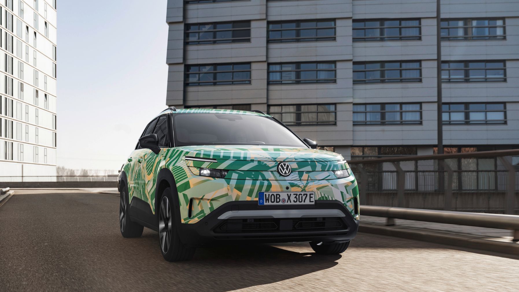 Ein ID. Cross von Volkswagen mit einem auffälligen grünen und gelben Camouflage-Design fährt auf einer städtischen Straße. Im Hintergrund sind moderne Gebäude zu sehen. Das Nummernschild des Autos beginnt mit "WOB".