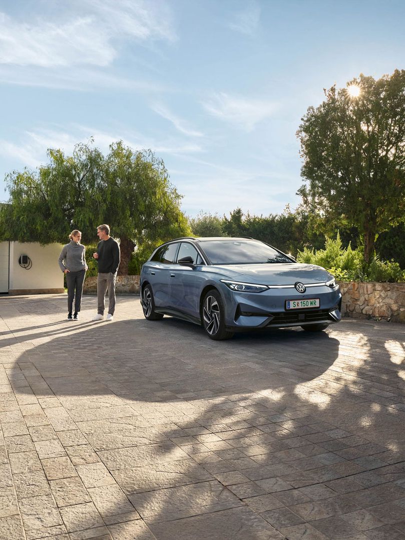 Der VW ID.7 Tourer ist vor Einfamillienhaus geparkt, eine Frau und ein Mann stehen neben ihm und unterhalten sich