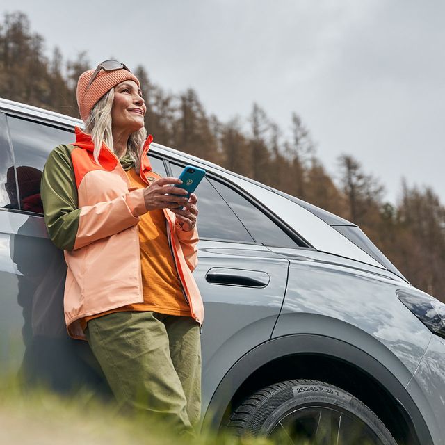 Eine Frau  mit Smartphone lehnt an einem Volkswagen und blickt in die Ferne