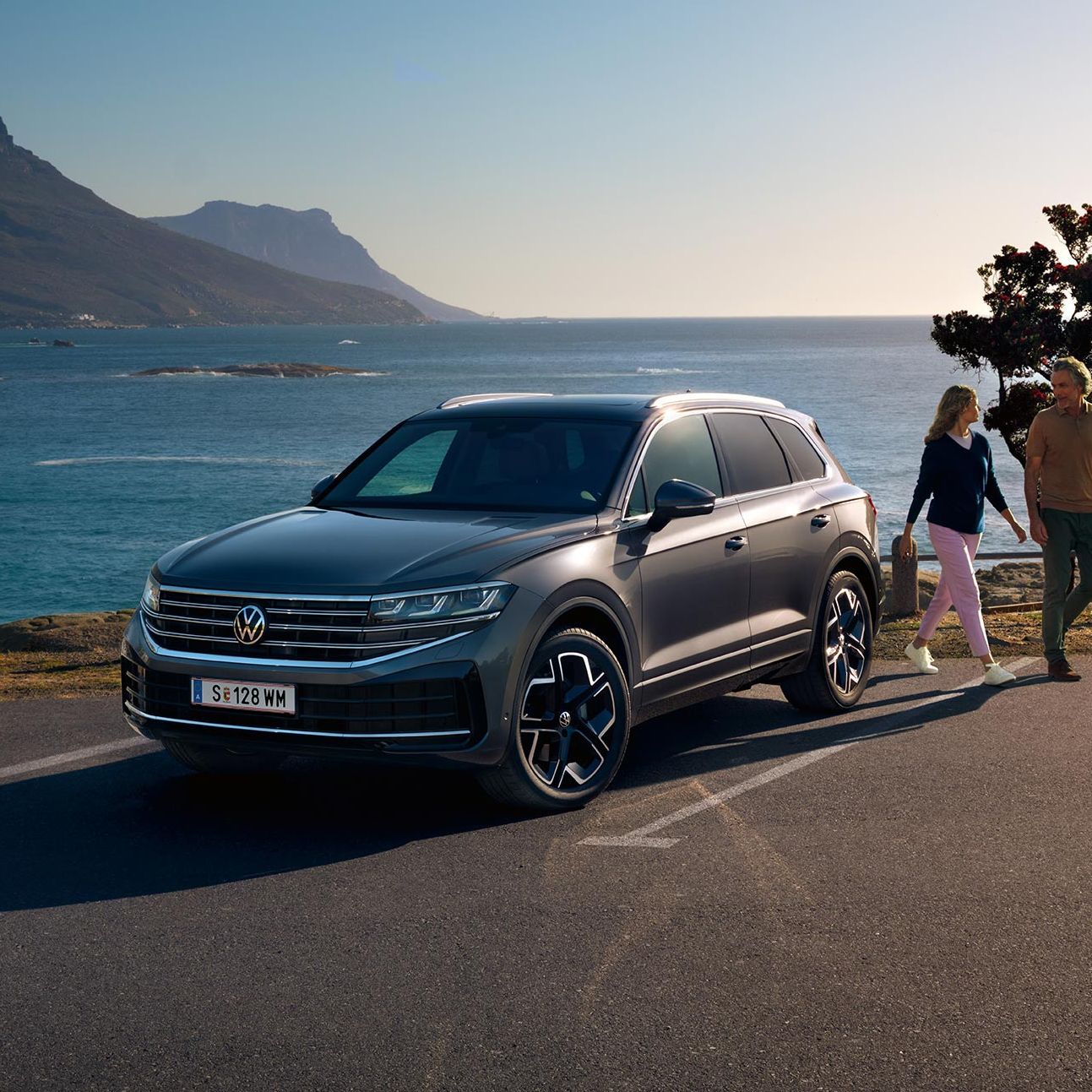 Der neue VW Touareg auf einem Parkplatz an der Küste, an ihm gehen eine Frau und ein Mann vorbei