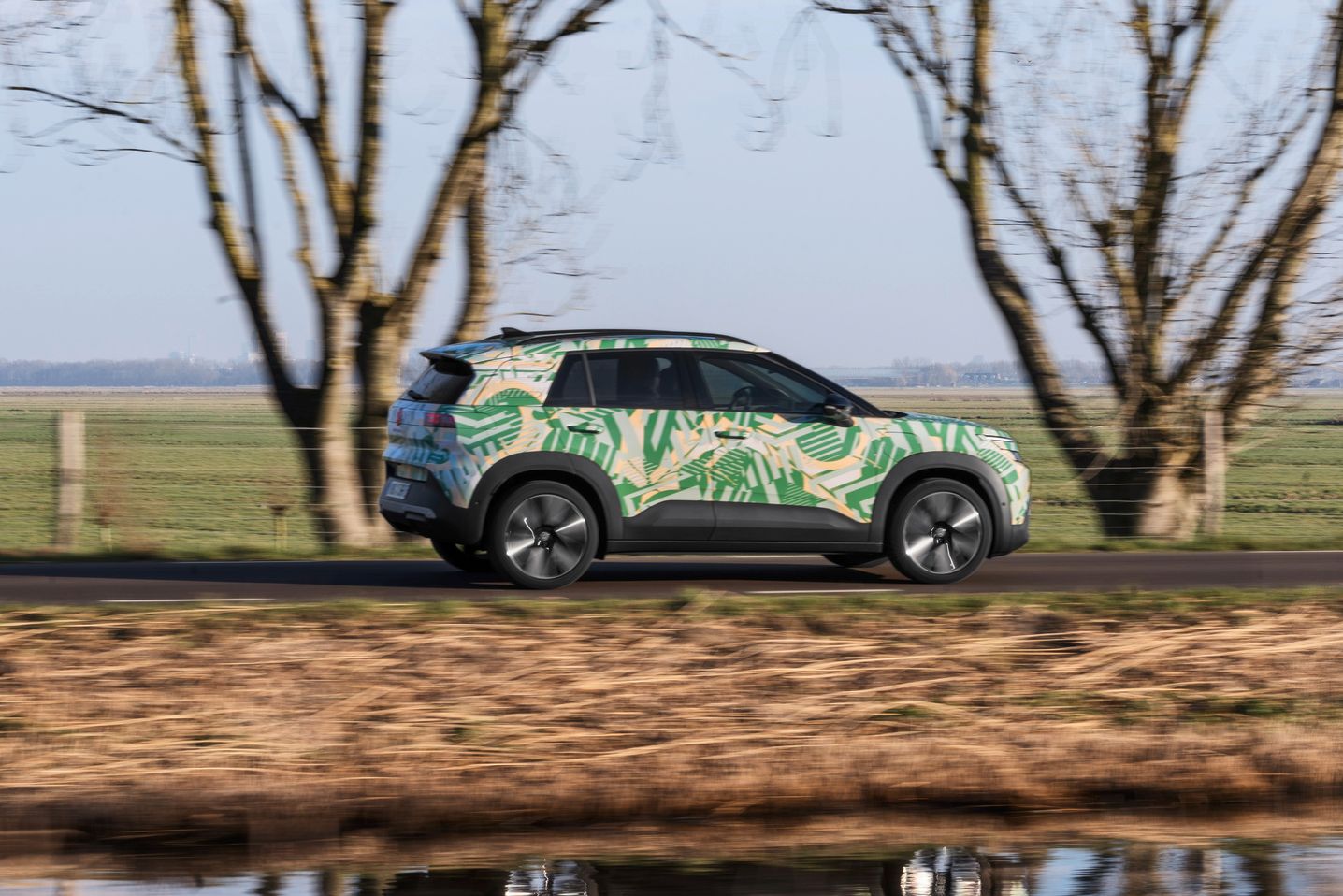 Ein SUV fährt auf einer ländlichen Straße vorbei an kahlen Bäumen und einem Wassergraben. Das Auto ist mit einem bunten, tropischen Muster bemalt. Im Hintergrund ist eine grüne Wiese und wolkenloser Himmel zu sehen.