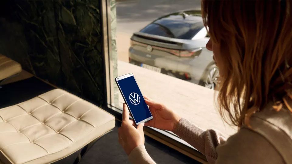 Eine Frau blickt auf ihr Smartphone, durch das Fenster ist ein VW ID. zu sehen