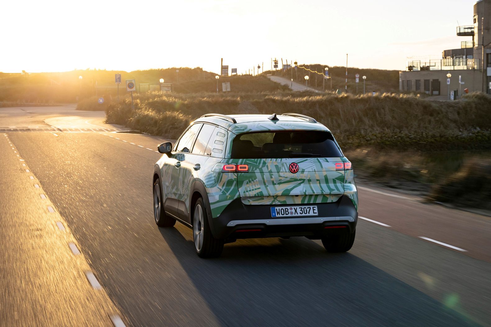 Ein Auto fährt auf einer Straße in einer Landschaft bei Sonnenuntergang. Das Fahrzeug hat ein grünes und weißes Design und ist ein elektrisches Modell. Im Hintergrund sind Gebäude und Hügel zu sehen.