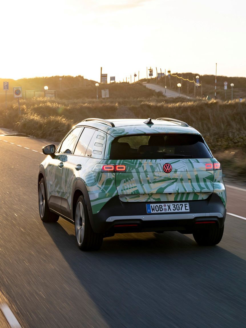 Ein Auto fährt auf einer Straße in einer Landschaft bei Sonnenuntergang. Das Fahrzeug hat ein grünes und weißes Design und ist ein elektrisches Modell. Im Hintergrund sind Gebäude und Hügel zu sehen.