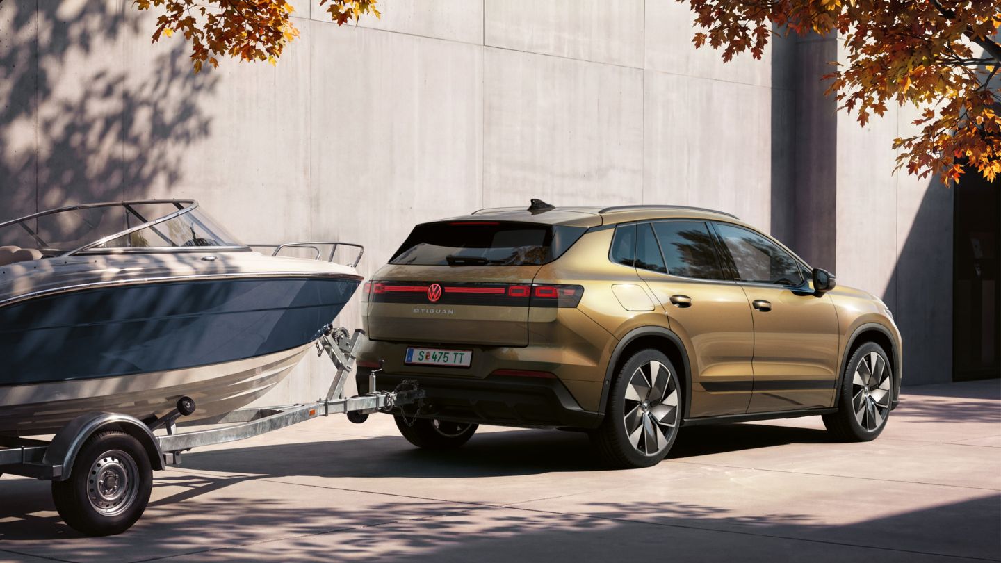 Ein oliv-bronzefarbener VW ID.4 Reskin parkt vor einem modernen Gebäude. Er zieht einen Anhänger mit Boot.