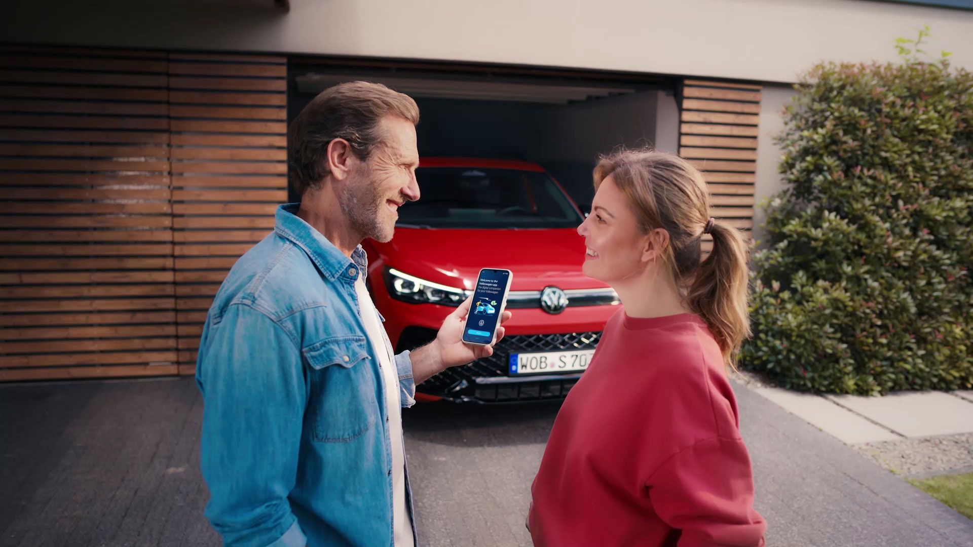 Ein Mann und eine Frau stehen vor einer Garage, in der ein rotes Auto (VW) geparkt ist. Der Mann zeigt der Frau etwas auf seinem Smartphone. Beide Personen haben unscharfe Gesichter.