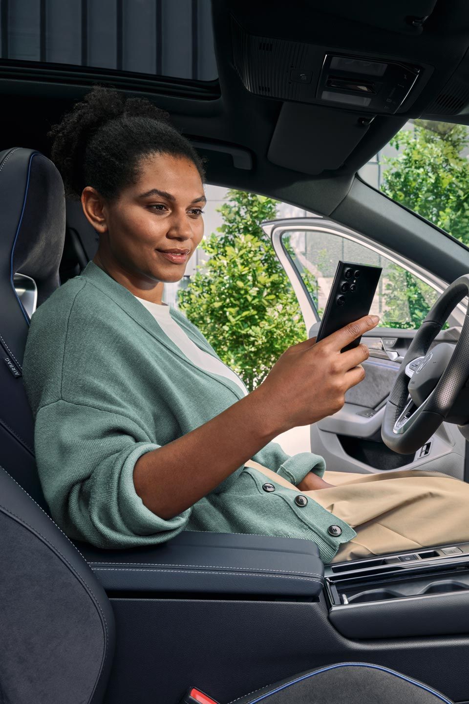 Eine Frau sitzt bei geöffneter Tür auf dem Fahrersitz des geparkten VW Passat und blickt auf ihr Smartphone.