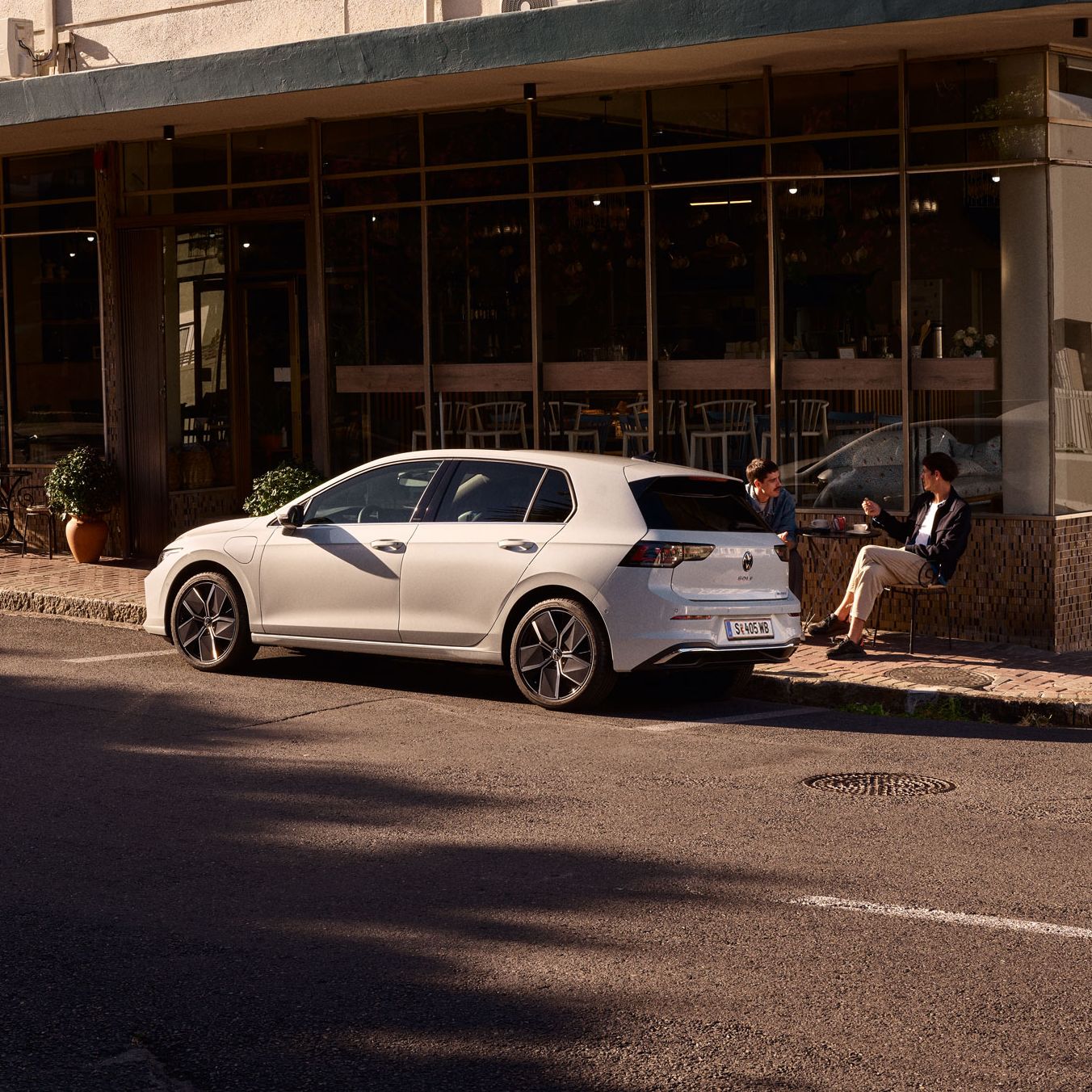 Zwei Männer sitzen vor einem Cafe und unterhalten sich. Vor ihnen ein neuer Golf 8.