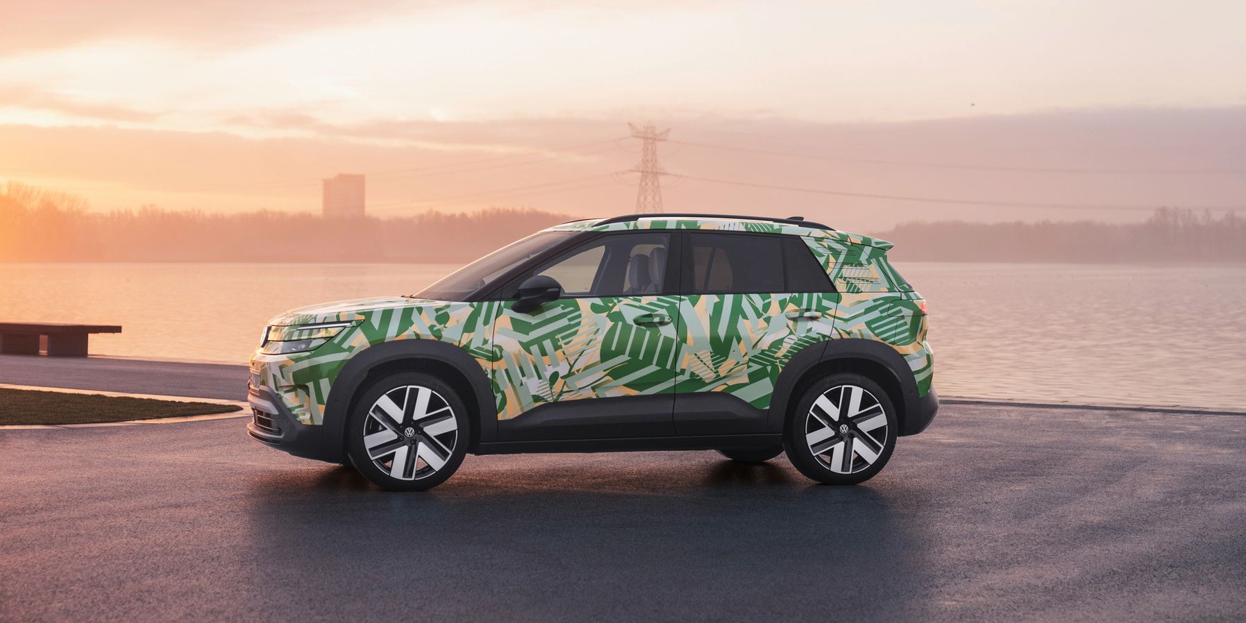 Ein SUV mit einer grünen und weißen Tarnlackierung steht auf einem asphaltierten Platz vor einem Gewässer bei Sonnenuntergang. Im Hintergrund sind Strommasten und Gebäude zu sehen. Der Himmel ist leicht bewölkt und in warmen Farben gehalten.