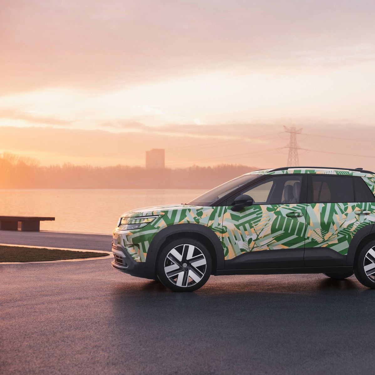 Ein SUV mit einer grünen und weißen Tarnlackierung steht auf einem asphaltierten Platz vor einem Gewässer bei Sonnenuntergang. Im Hintergrund sind Strommasten und Gebäude zu sehen. Der Himmel ist leicht bewölkt und in warmen Farben gehalten.