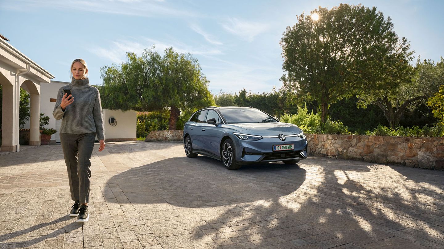 Der VW ID.7 Tourer ist vor Einfamilienhaus geparkt, eine Frau geht von ihm weg und blickt auf ihr Smartphone