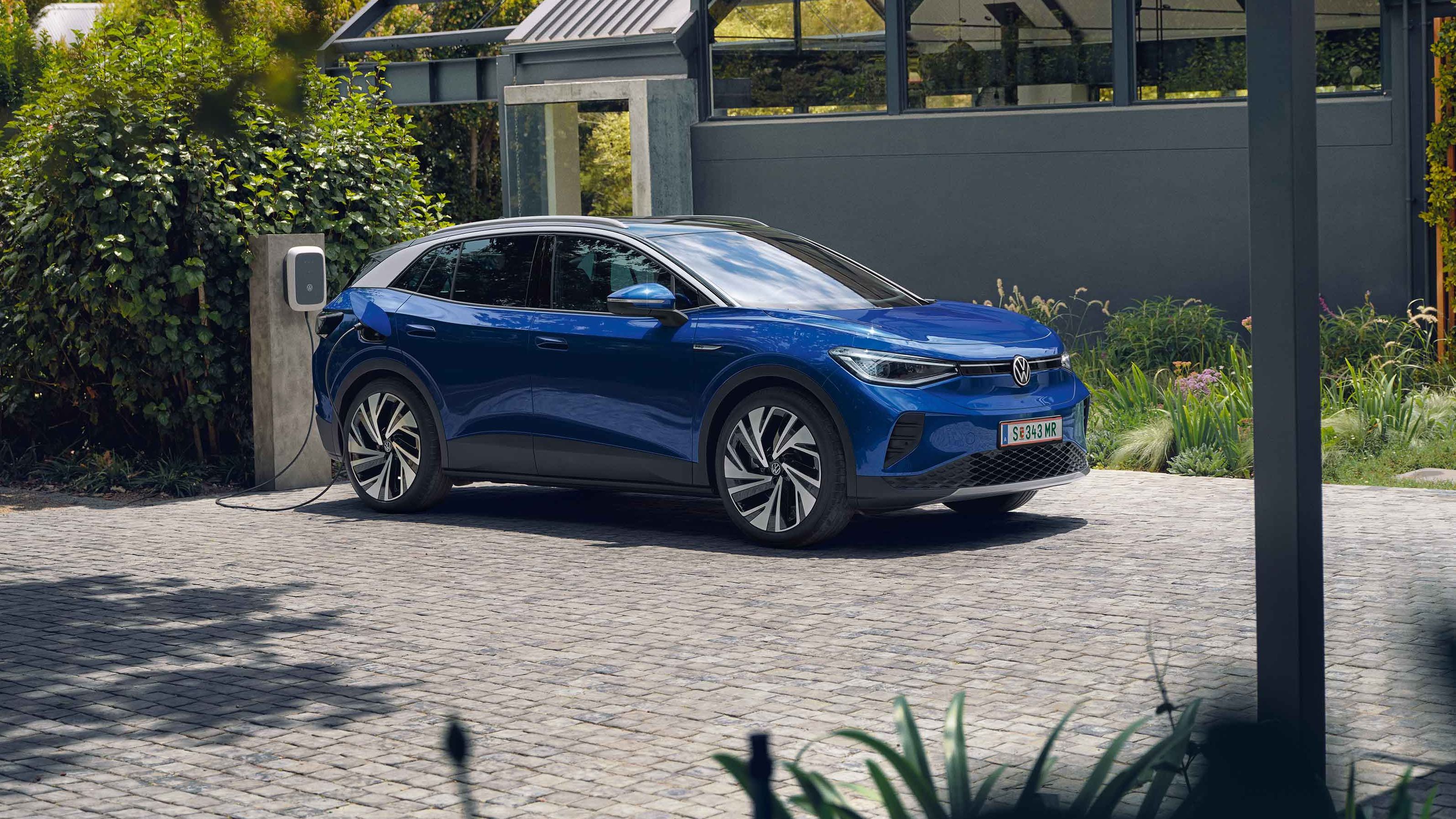 Ein blauer VW ID.4 parkt seitlich vor einem grauen, modernen Haus und lädt an der ID. Charger Wallbox. Im Hintergrund viel grün.