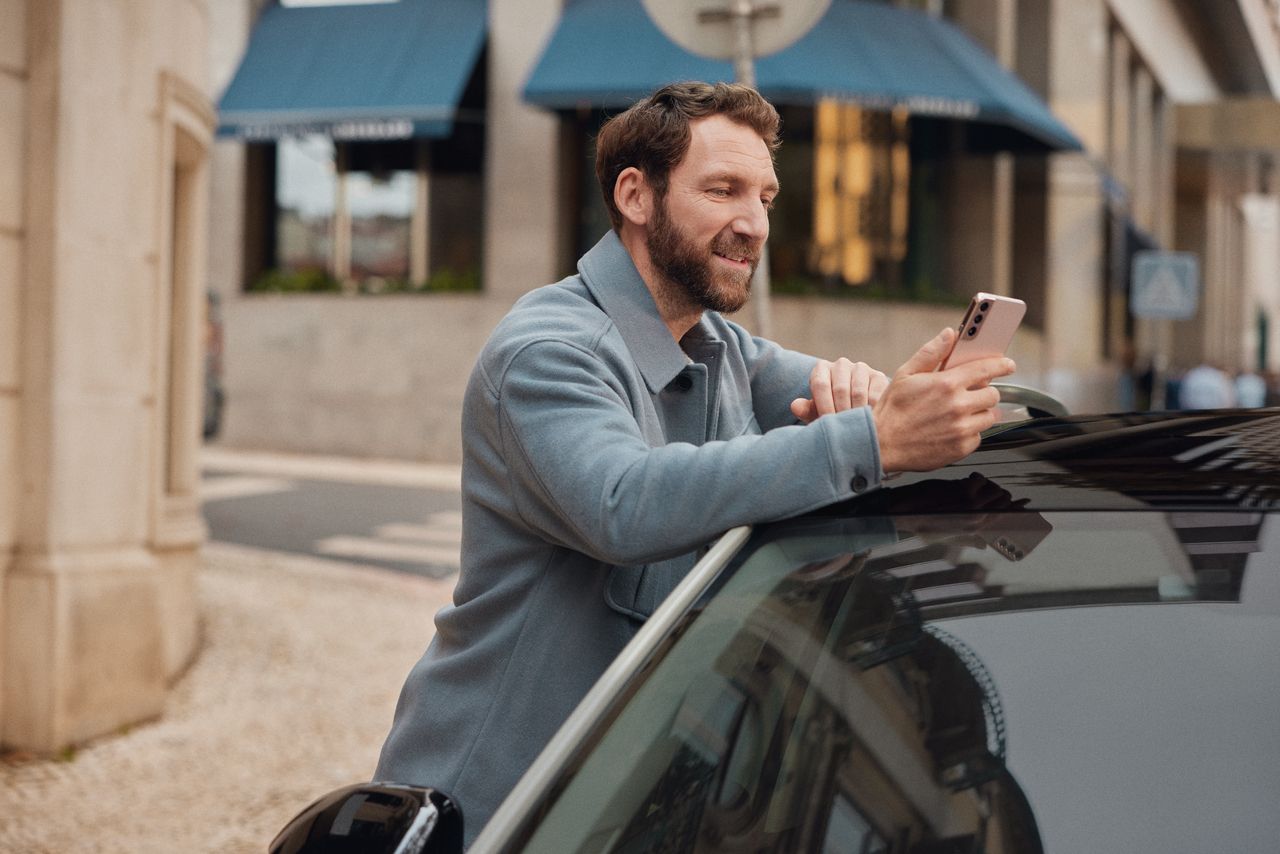 Mann lehnt an einem Volkswagen und schaut auf sein Mobiltelefon