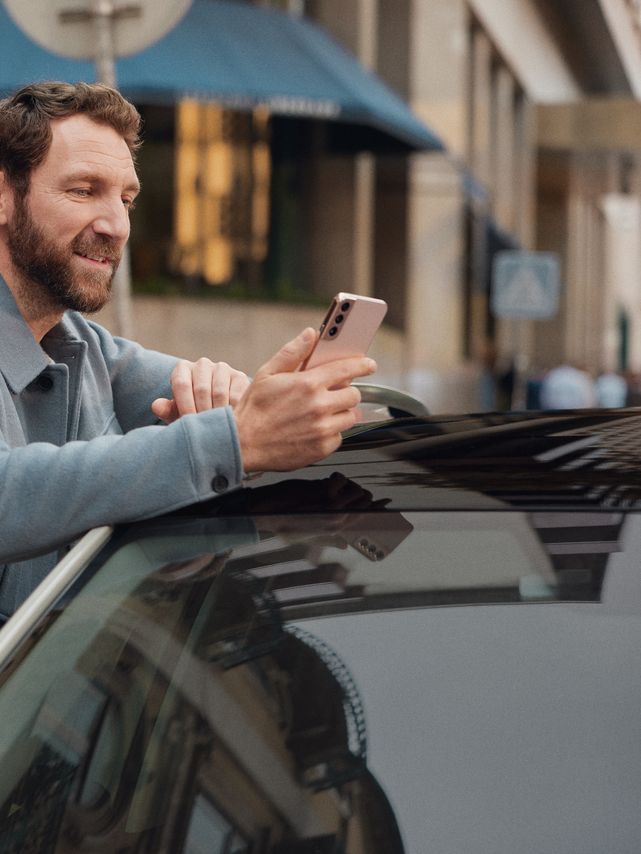 Mann lehnt an einem Volkswagen und schaut auf sein Mobiltelefon