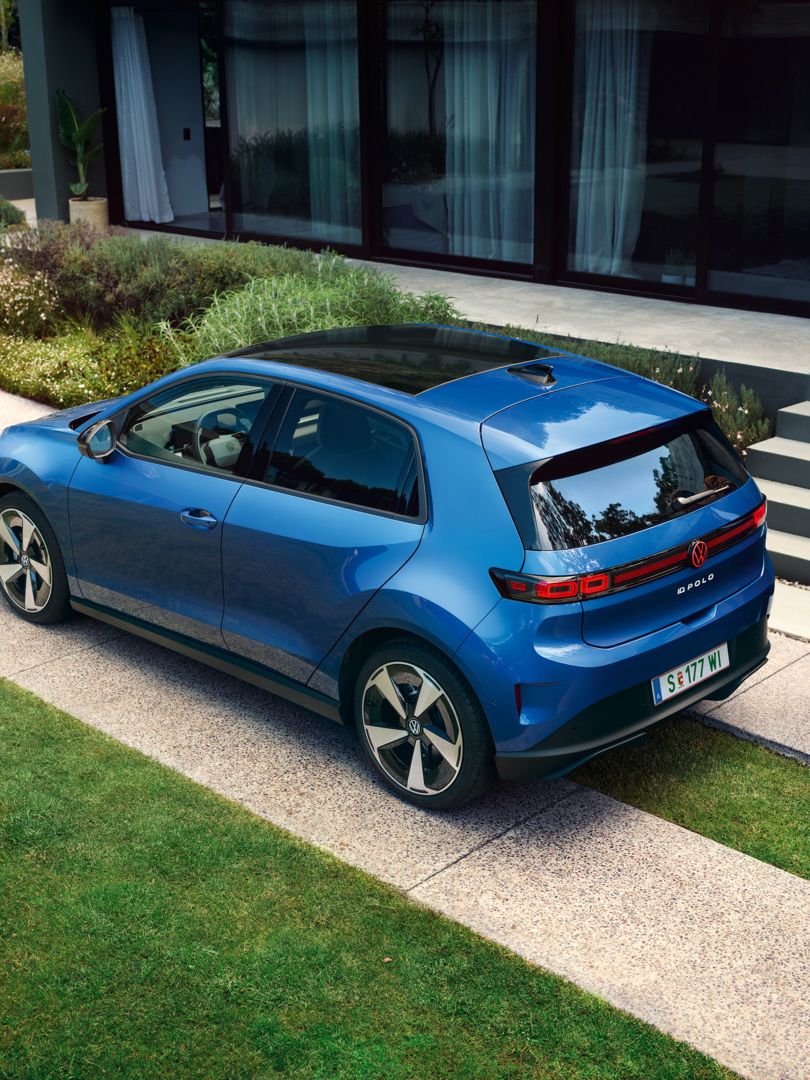 Ein blauer VW ID. Polo Style parkt vor einem begrünten Wohnhaus, ein Mann geht hinter dem Fahrzeug entlang.